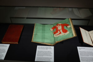 Books displayed in cabinet including sample of red and white knitting with instruction
