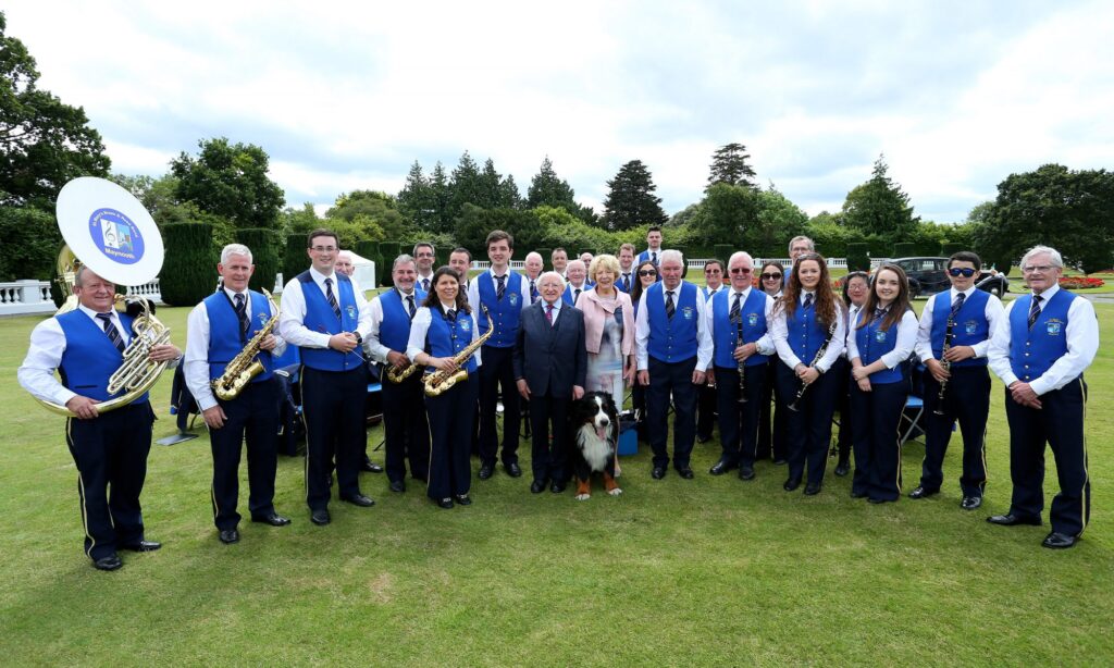 Brass band wearing blue waistcoats posing with President Michael D Higgins, wife Sabina and pet dog Bród